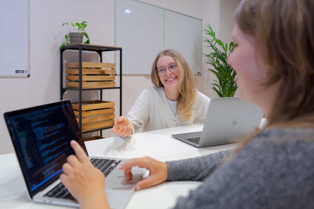 Zwei Frauen arbeiten gemeinsam an Laptops, eine mit Code auf dem Bildschirm, während die andere lächelt und zuhört.