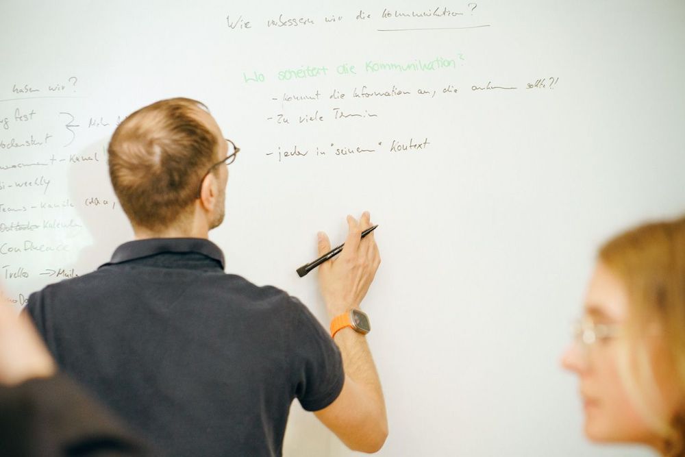 Ein Mann schreibt während eines Workshops Ideen auf ein Whiteboard, während Kolleginnen aufmerksam zuhören.